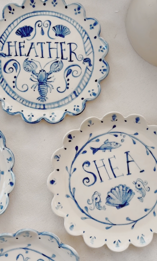 A close-up of decorative ceramic plates with scalloped edges, featuring blue hand-painted sea-themed illustrations and names including 'HEATHER' and 'SHEA'.